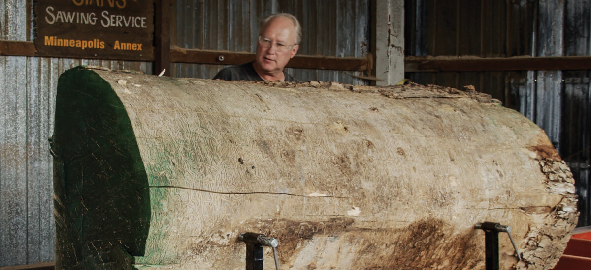 Person standing behind an oversized reclaimed wood log in a workshop, showing natural bark texture and cut ends.