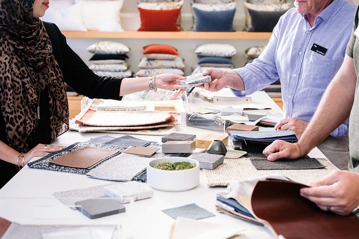 Hands exchanging material samples over a table with fabric, leather, and finish swatches.