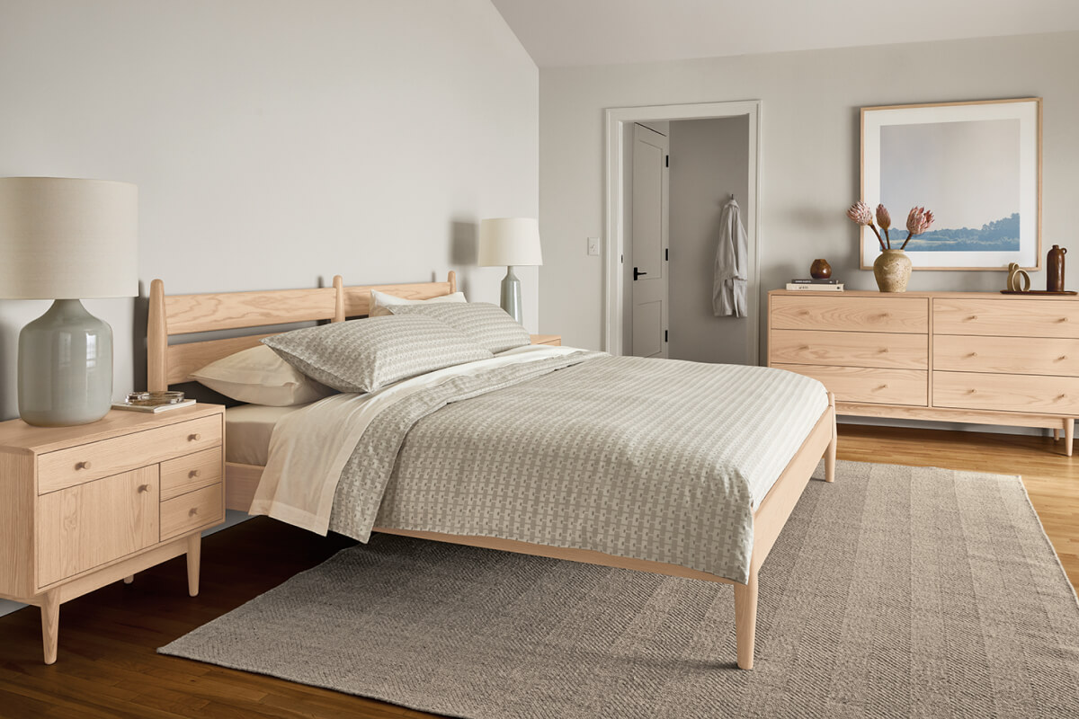 Grove bed in light wood with grey bedding, surrounded by matching nightstands and dresser with table lamp and decor.
