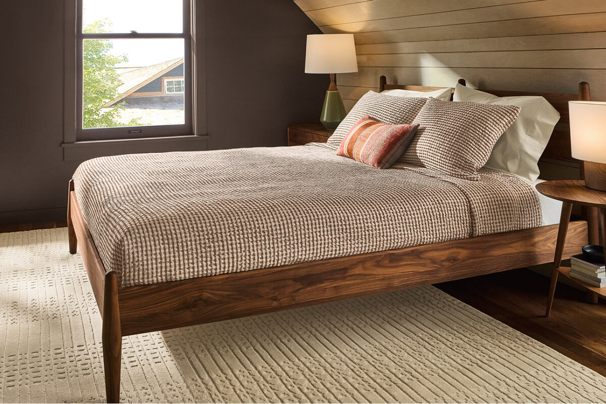 Grove bed in walnut with textured bedding and pillows in a bedroom with wood panel wall and window.