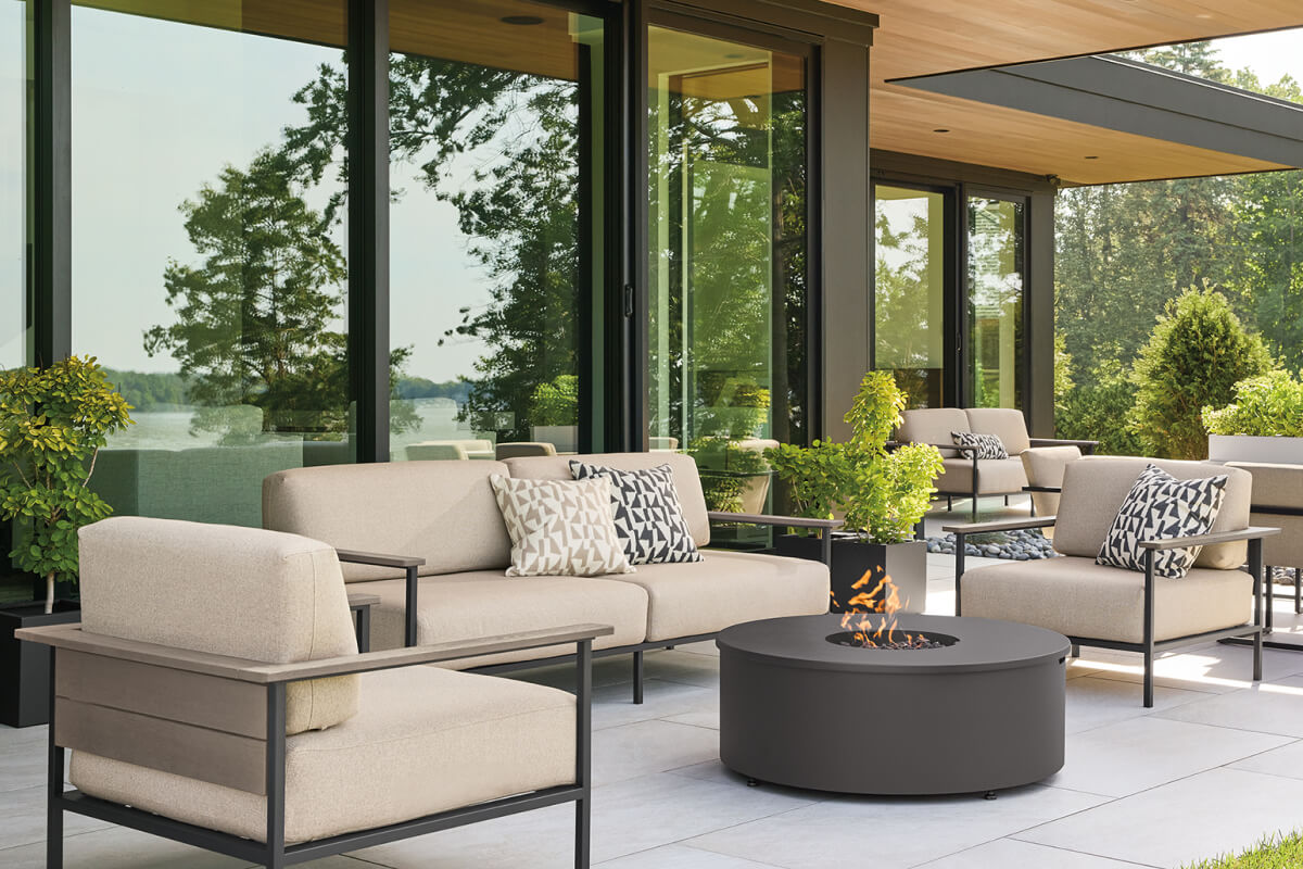 Marin outdoor sofas and chairs with grey frames and light cushions around a round fire table on a patio.