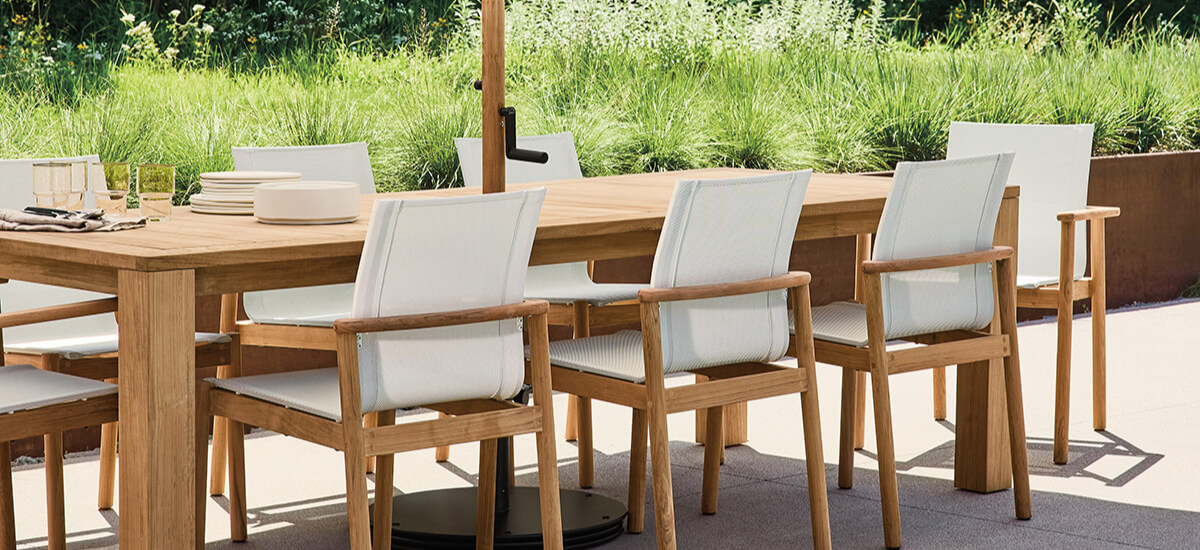 Teak Capri outdoor dining table with Peri chairs and light cushions arranged on a patio with greenery in the background.