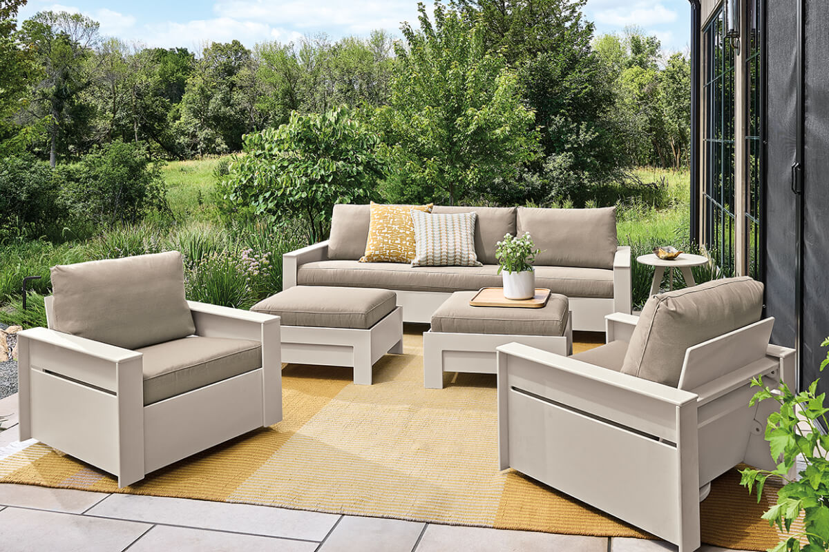 Rayo outdoor sofa and swivel chairs with beige cushions on a patio with matching ottomans and rug.