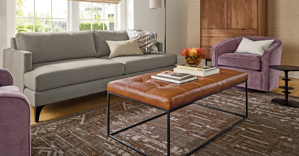 Amira rug with a geometric brown and ivory pattern beneath a grey sofa, leather ottoman and accent chairs.