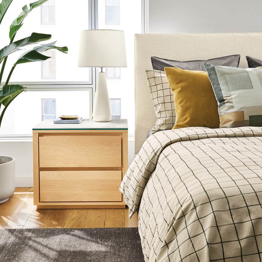 Two-drawer Arro nightstand in maple with white stone top, styled next to a cream bed with patterned linens.