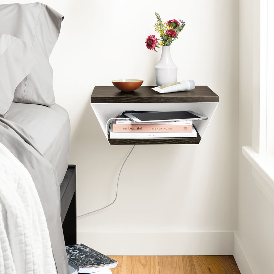 Alta wall shelf with wood top and open storage, holding books, a tablet and vase beside a bed with white linens.