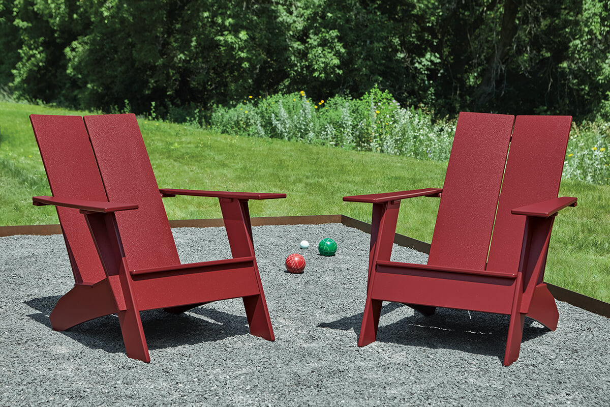 Pair of Emmet outdoor chairs in red finish, positioned on a gravel bocce court with lawn and trees beyond.