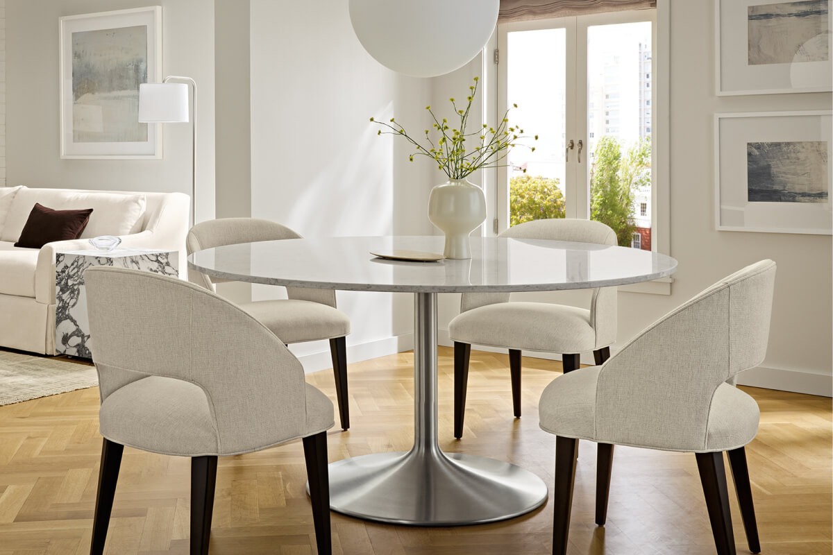 Aria table with a round white stone top and upholstered Esme chairs, styled with a vase centerpiece and pendant lighting.