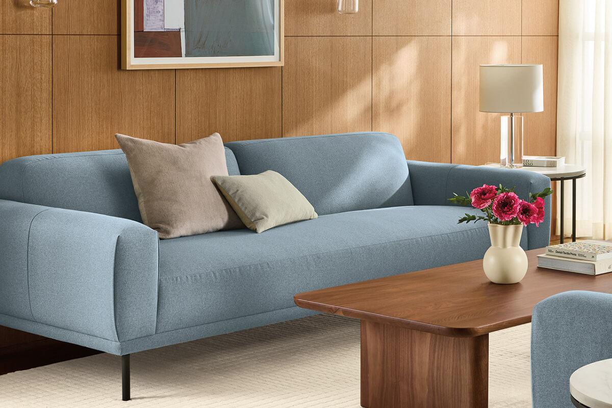 Light blue Ali sofa with neutral pillows, paired with a walnut coffee table and floral arrangement.