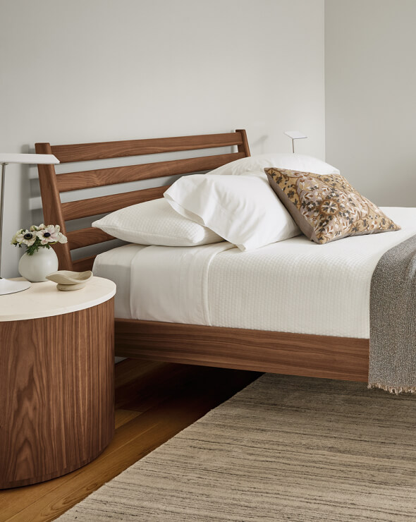 Silas bed in walnut with a slatted headboard, white bedding, and a round walnut nightstand.