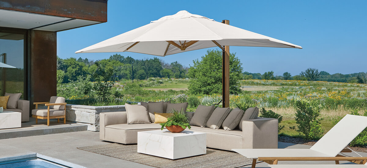 Beige outdoor sectional and white Cumulo umbrella on a patio beside a pool overlooking a green landscape.