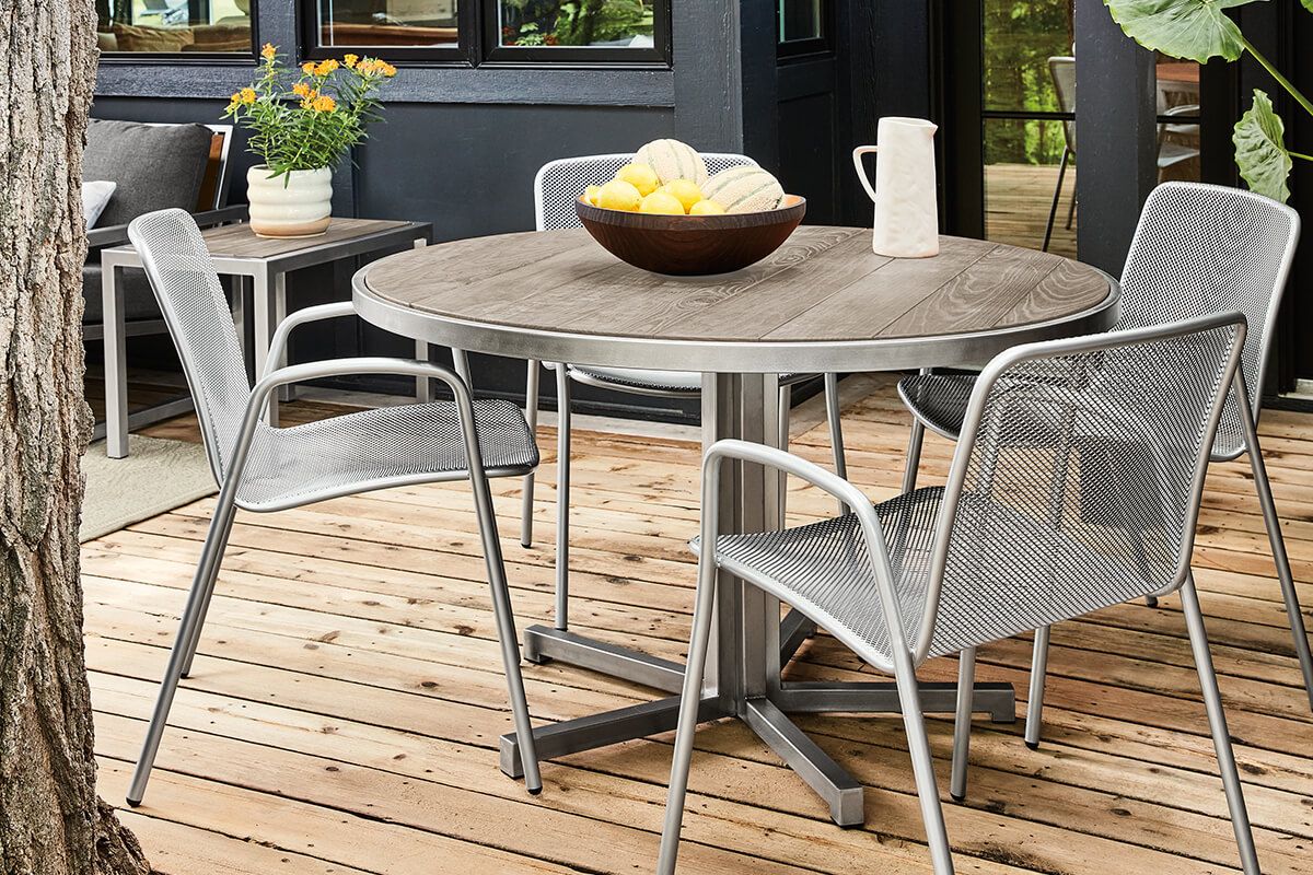 Montego round dining table with a solid wood top and brushed metal pedestal base, surrounded by four mesh outdoor chairs.