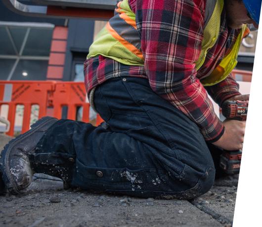 Image of a man in a safety vest and helmet crouched on the ground wearing dark work boots and using a drill.