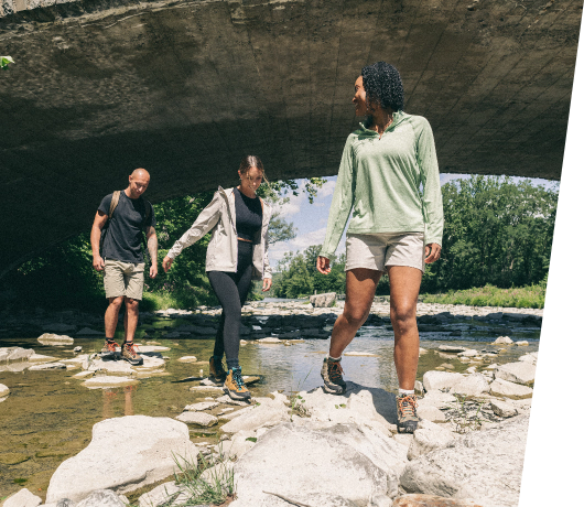 Three people in the summer time crossing a stream under a concrete bridge, wearing hiking boots.