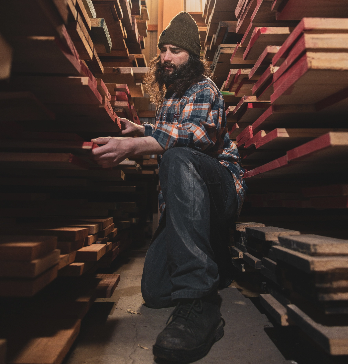 A man in a beanie, jeans, and a plaid shirt chooses from among planks of wood stored on the shelves surrounding him.