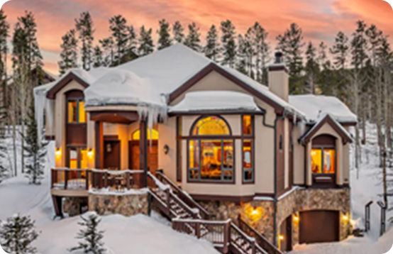 Snow-covered mountain chalet with large windows and glowing lights under a pink sunset sky.