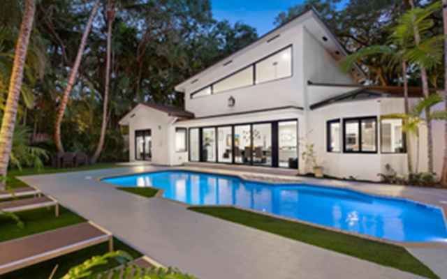 White modern home with sleek design, glowing pool, and lounge chairs set among tall palm trees at dusk.