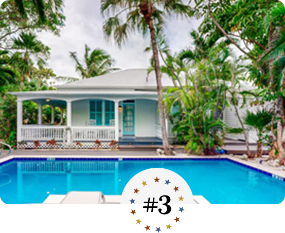 Charming coastal cottage with wraparound porch, lush palms, and inviting pool.