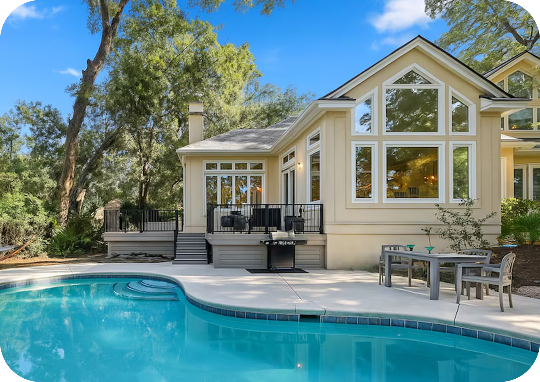 Backyard pool beside a light‑colored home with large windows and surrounding trees.
