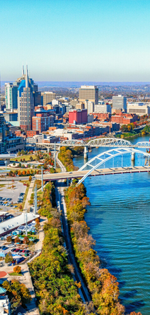 Nashville’s skyline rises above the Cumberland River, with a white arched bridge in the foreground.