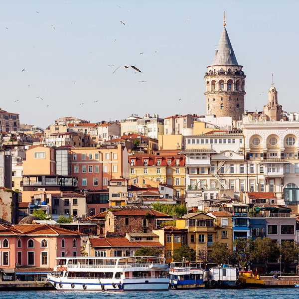 Istanbul waterfront with a ferry on the water, colorful hillside buildings, seagulls in the sky, and the Galata Tower rising above the city skyline.