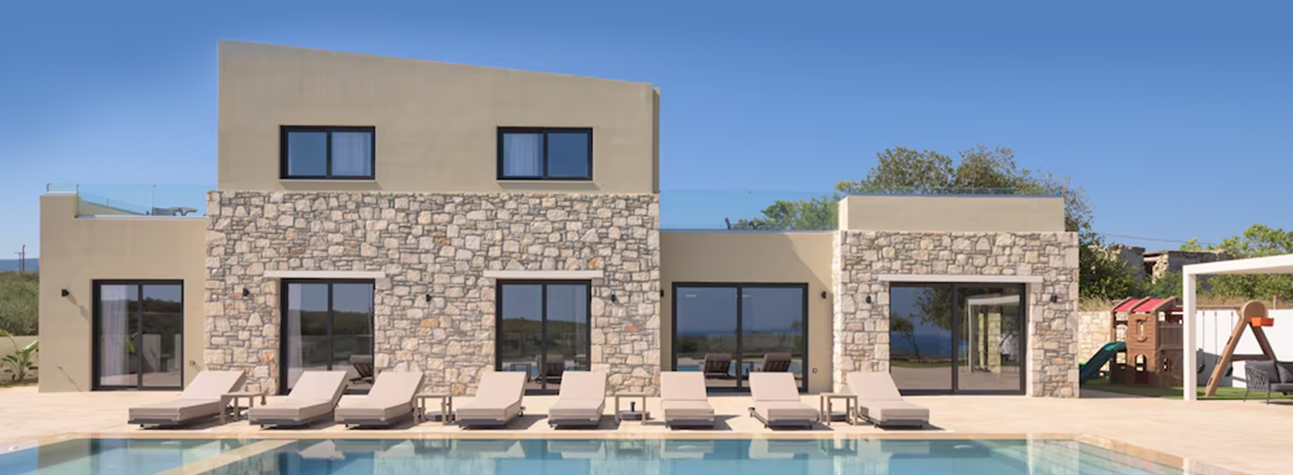 Stone villa with large windows, lounge chairs, and a pool.