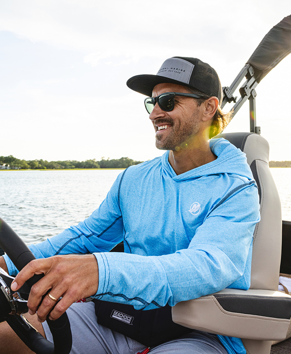 A man wearing a light blue long-sleeved shirt, sunglasses, and a baseball cap smiles as he steers a boat.