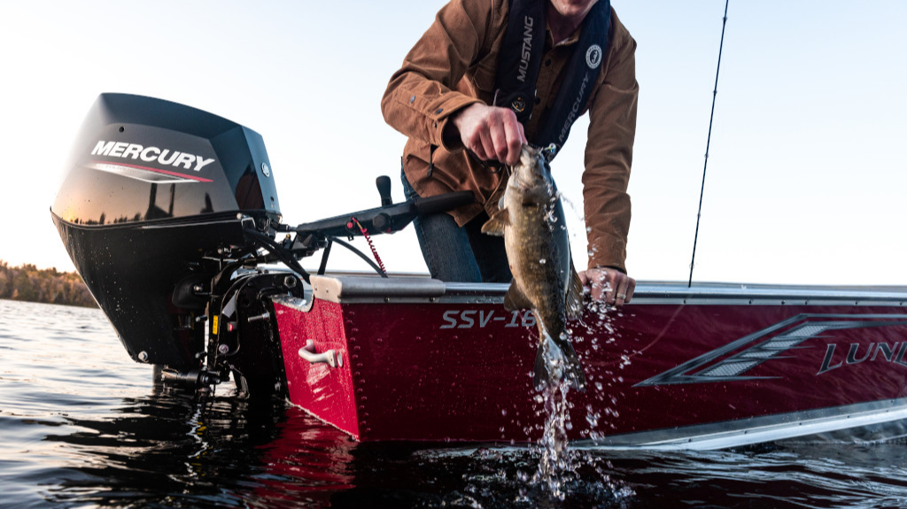 A fisherman in a Lund boat powered by a Mercury outboard motor pulls a smallmouth bass from the water.
