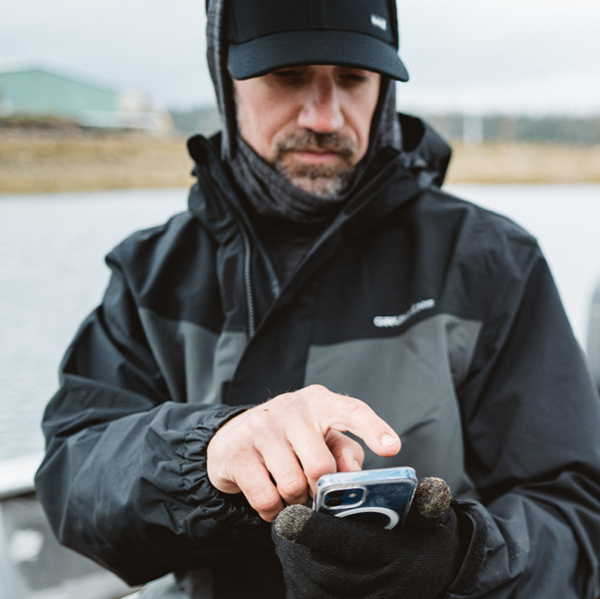 A man wearing a black jacket and gloves uses his phone on a boat.