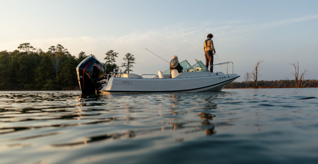 Two people fishing from a small motorboat on a calm lake at sunset.