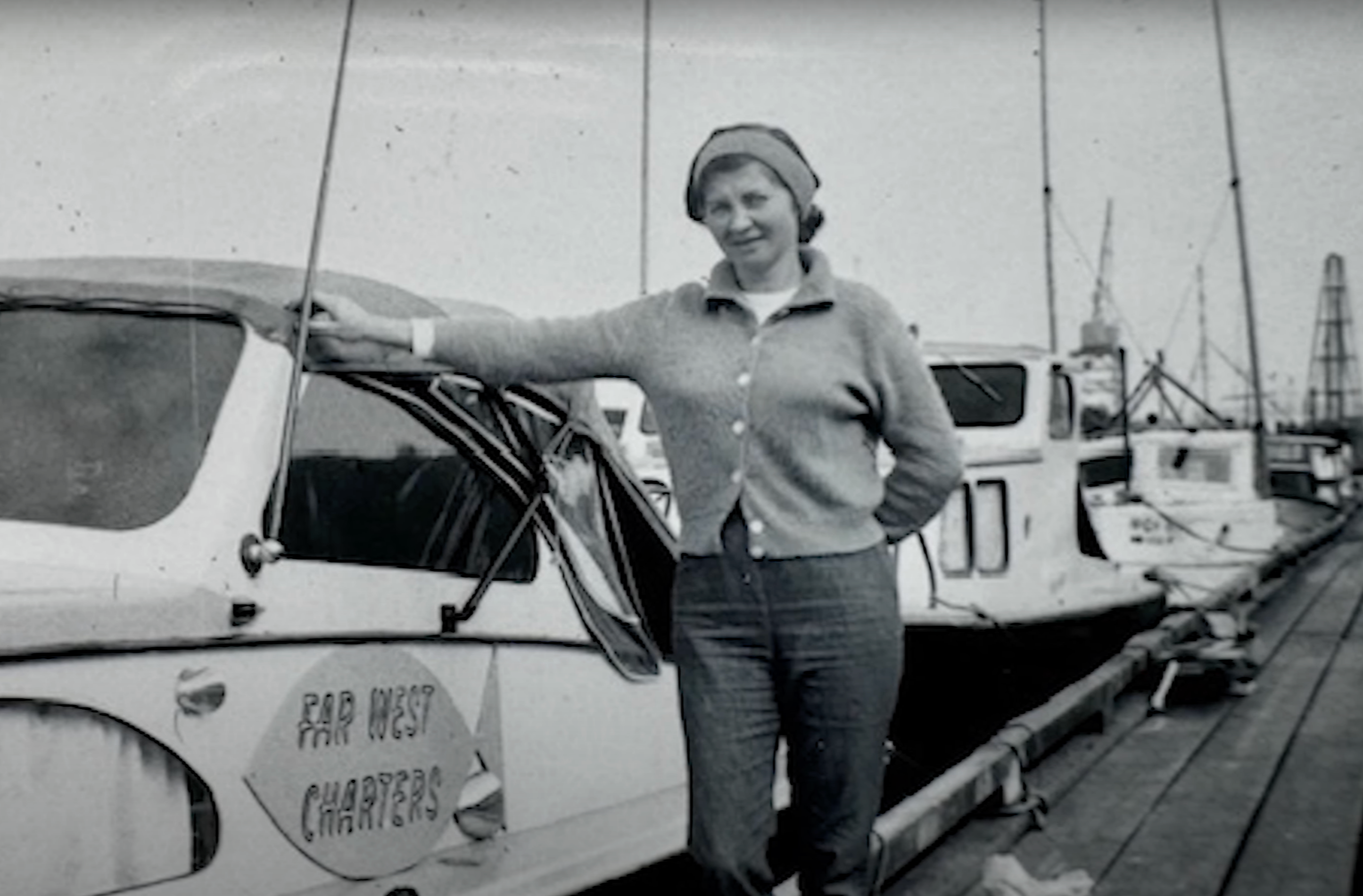 A black and white photo of a woman standing next to a boat with a sign that reads "Far West Charters". She's wearing a cardigan, pants, and a hat.
