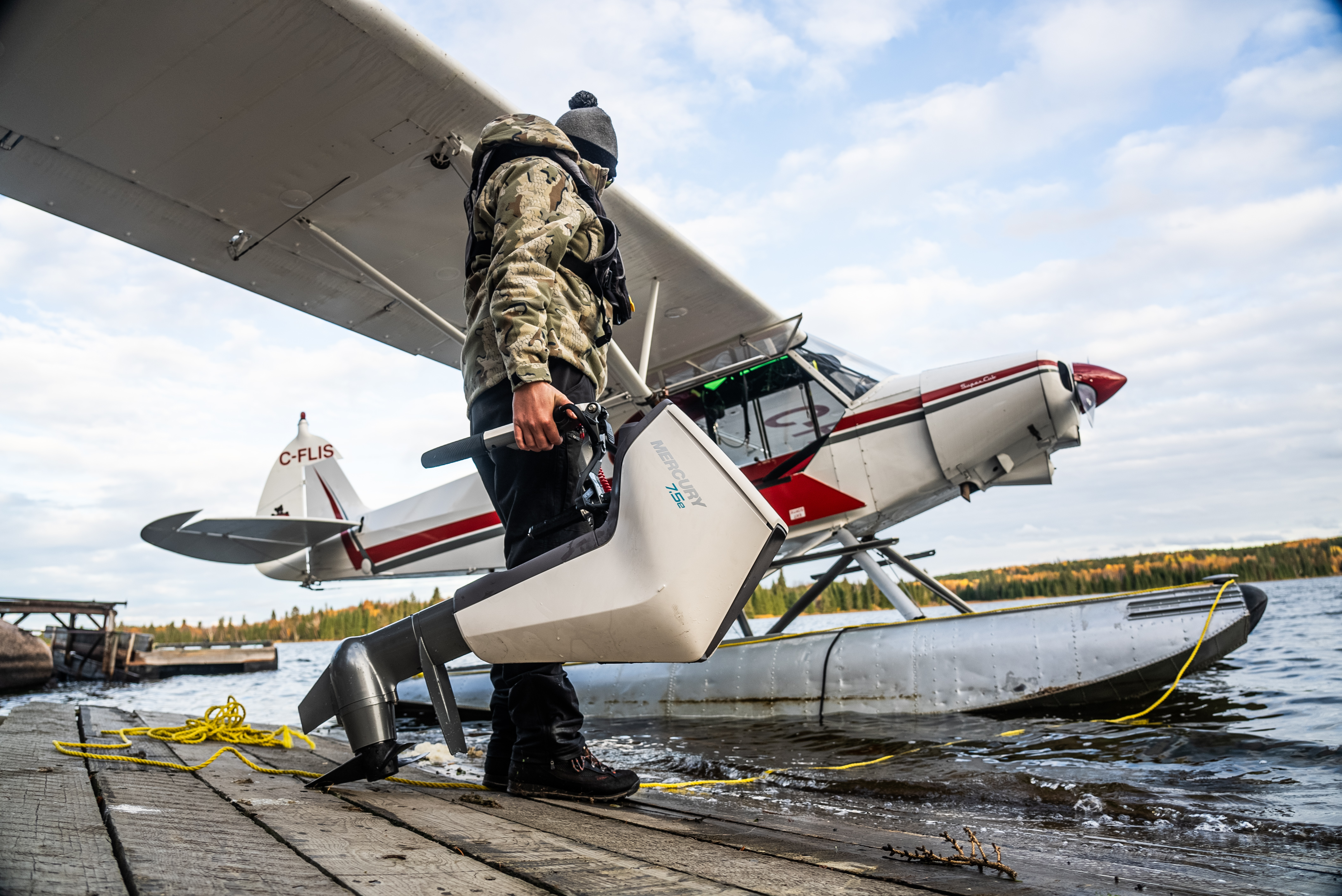 Someone in camouflage and black pants carries an outboard motor towards a plane waiting on a lake.