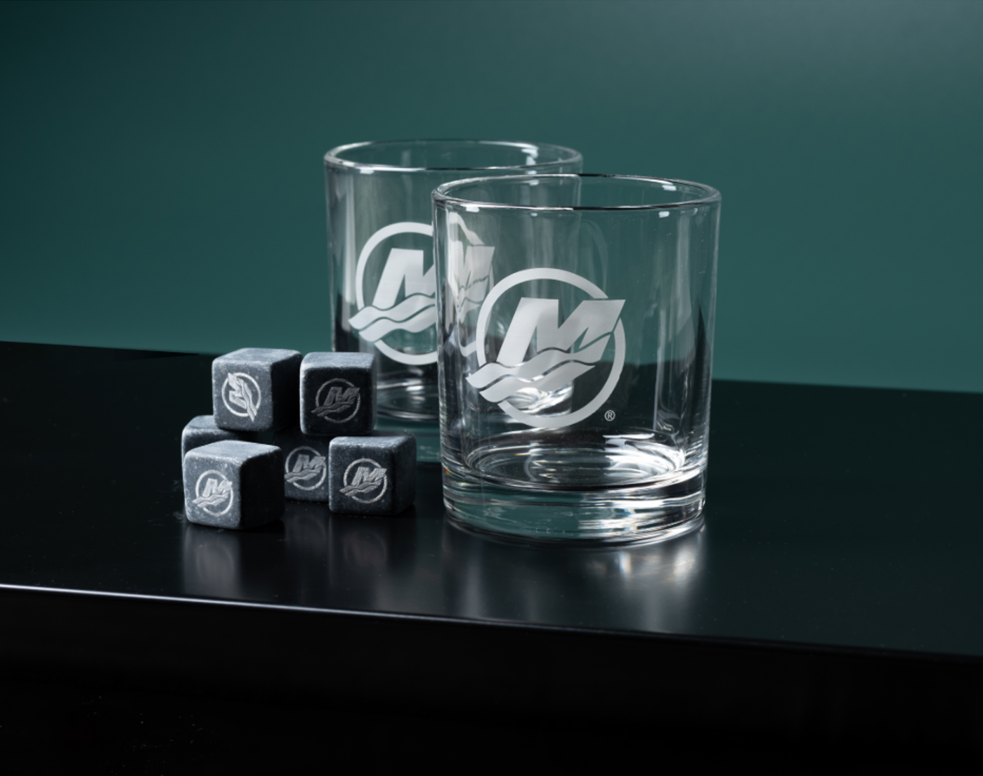Two clear whiskey glasses with a frosted "M" logo and a pile of whiskey stones on a dark surface.