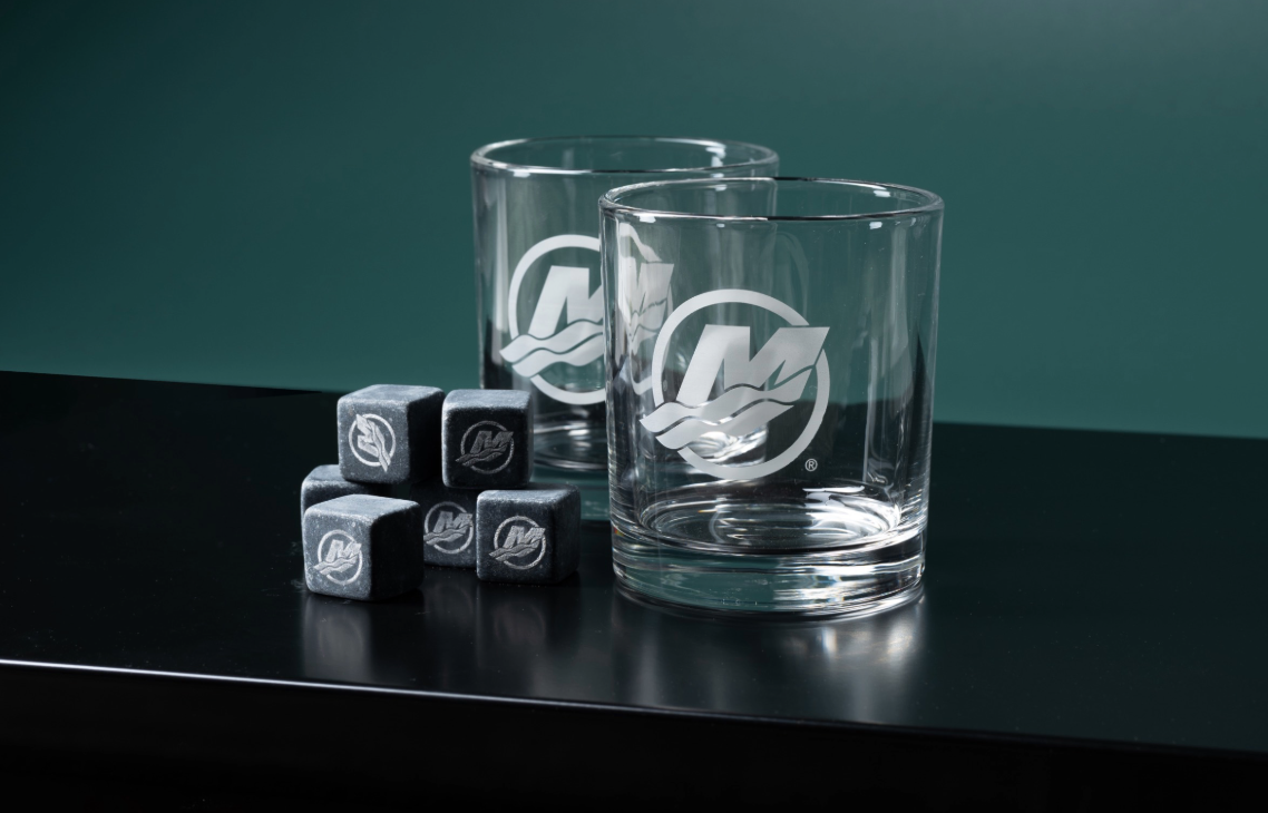 A pair of empty lowball glasses and matching whiskey stones, all featuring a stylized "M" logo.
