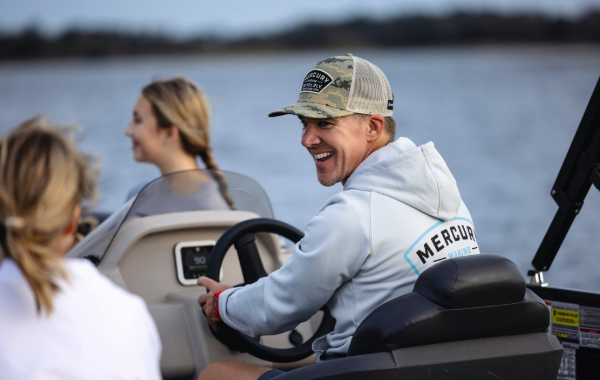 A man steering a boat on a lake, smiling back at his passengers.