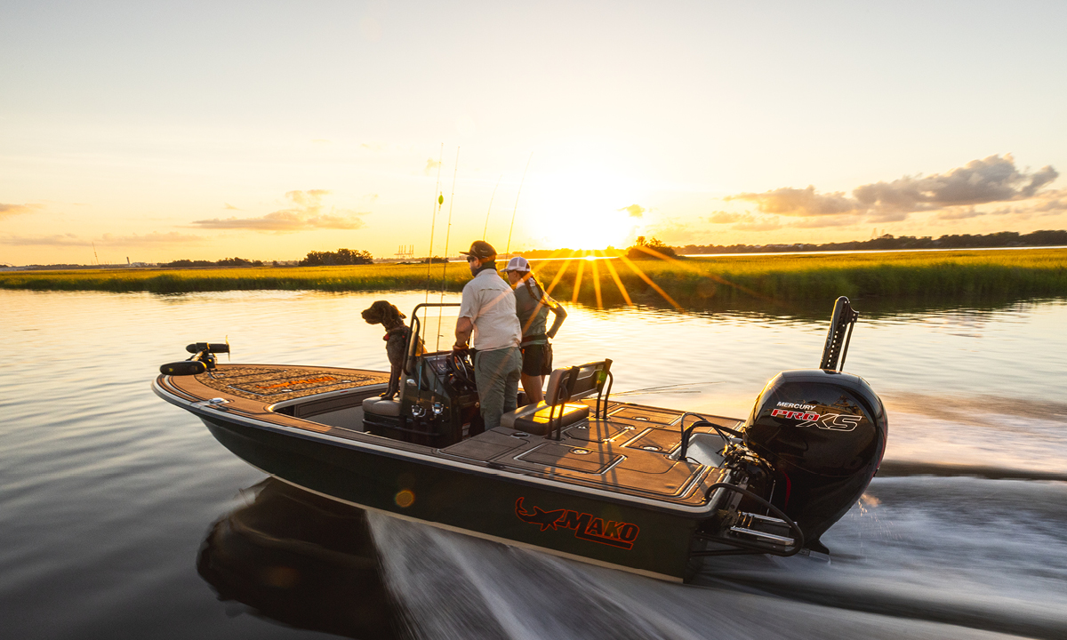 A man, a woman, and a dog are on a Mako boat with a Mercury Pro XS motor, enjoying a sunset cruise on a calm river.