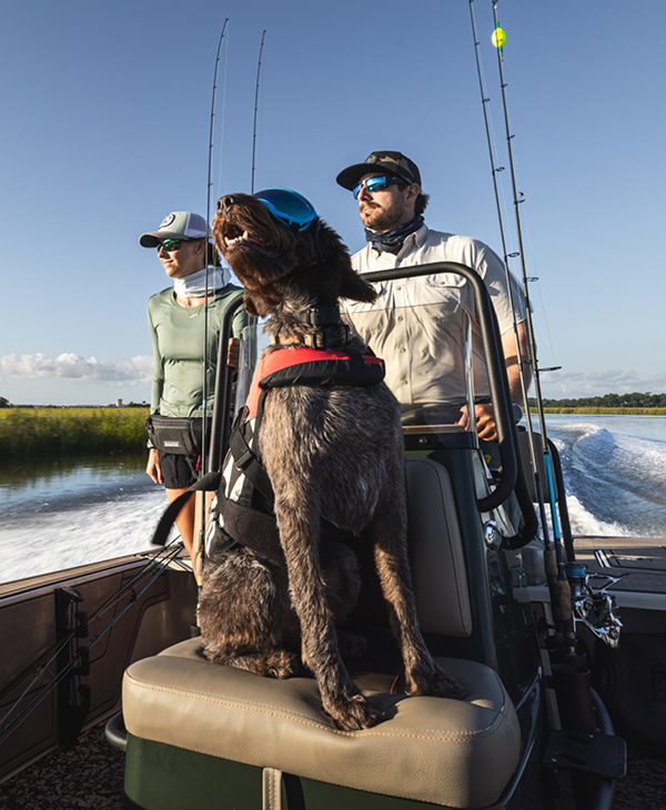A dog wearing a life vest sits on a boat seat between a man and a woman, enjoying a sunny day out fishing.