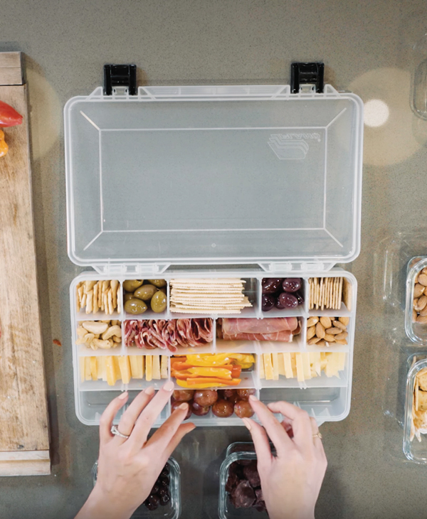 Overhead view of hands arranging a snack box filled with cheese, crackers, olives, cured meats, nuts, peppers, and grapes.