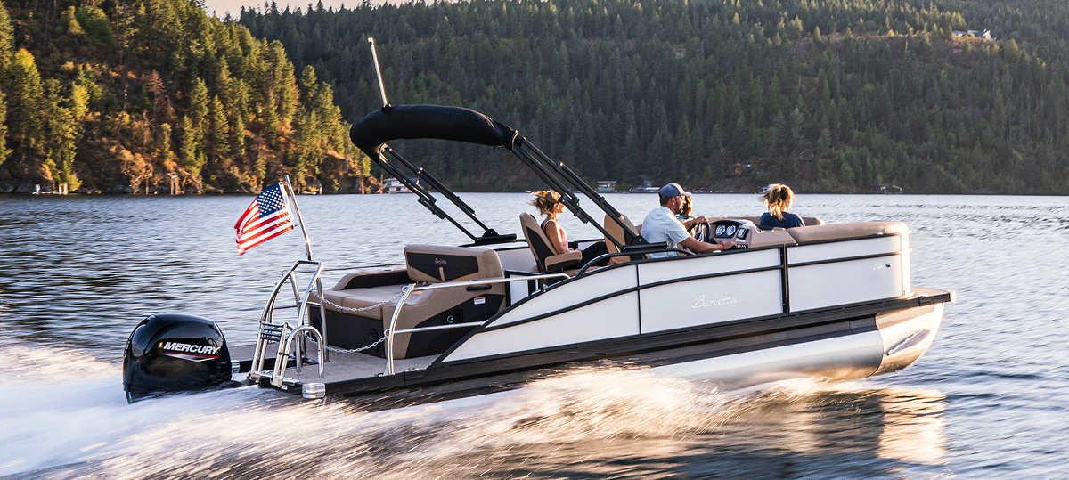 A Barletta pontoon boat speeds across a lake. A family of four enjoys the ride, with the driver at the helm. An American flag flies from the boat's railing.