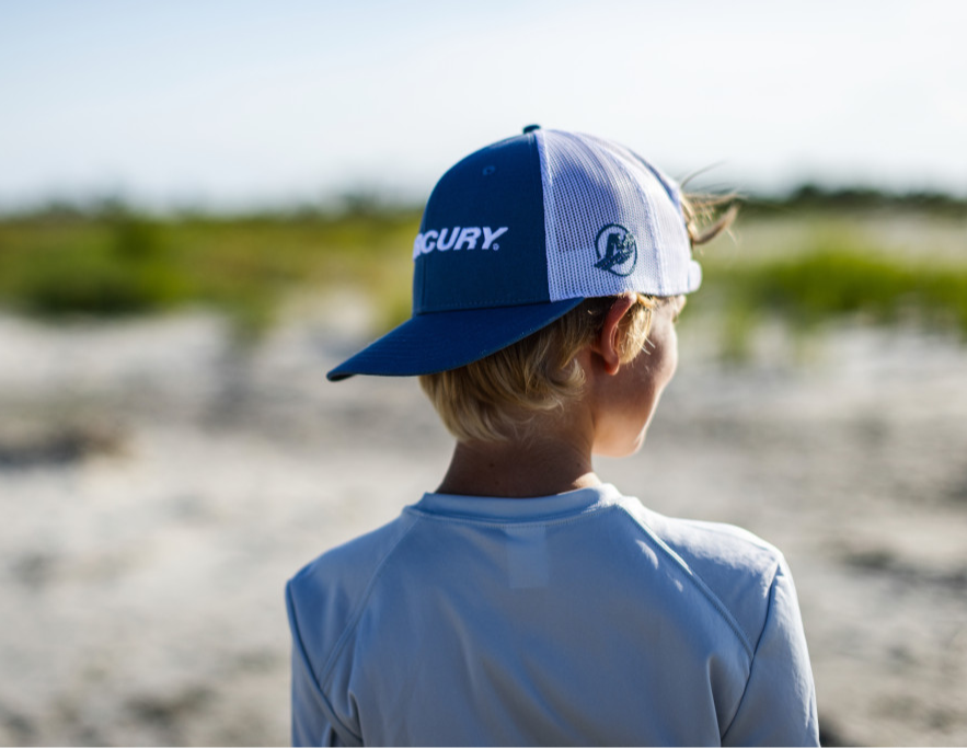 A blonde-haired person in a Mercury cap looking into the distance across a sandy beach or field.