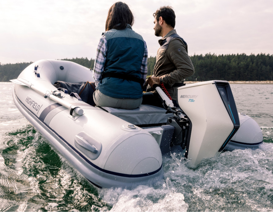 Two people ride in a Highfield inflatable boat powered by a Mercury 7.5e outboard motor, creating a wake on the water.