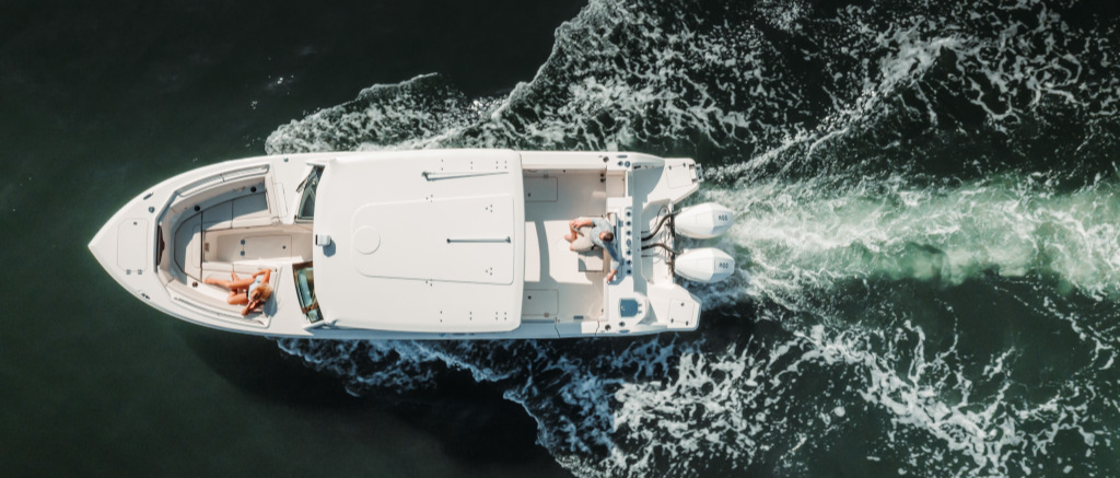Aerial view of a white boat with two people on board, cruising through dark water and leaving a foamy wake.