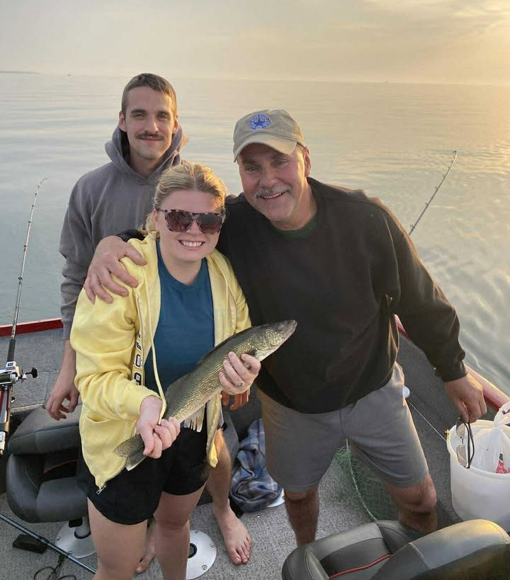 A smiling woman in sunglasses, a yellow jacket, and black shorts holds a fish on a boat, with two men standing beside her.