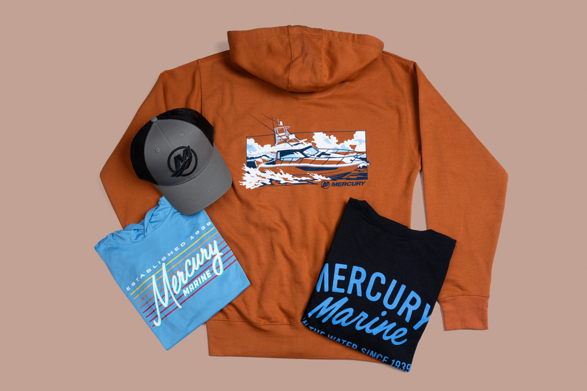 An orange hoodie with a boat graphic on the back, a blue t-shirt with a striped 'Mercury Marine' logo, a black t-shirt with a blue 'Mercury Marine' logo, and a grey cap with a black 'M' logo.