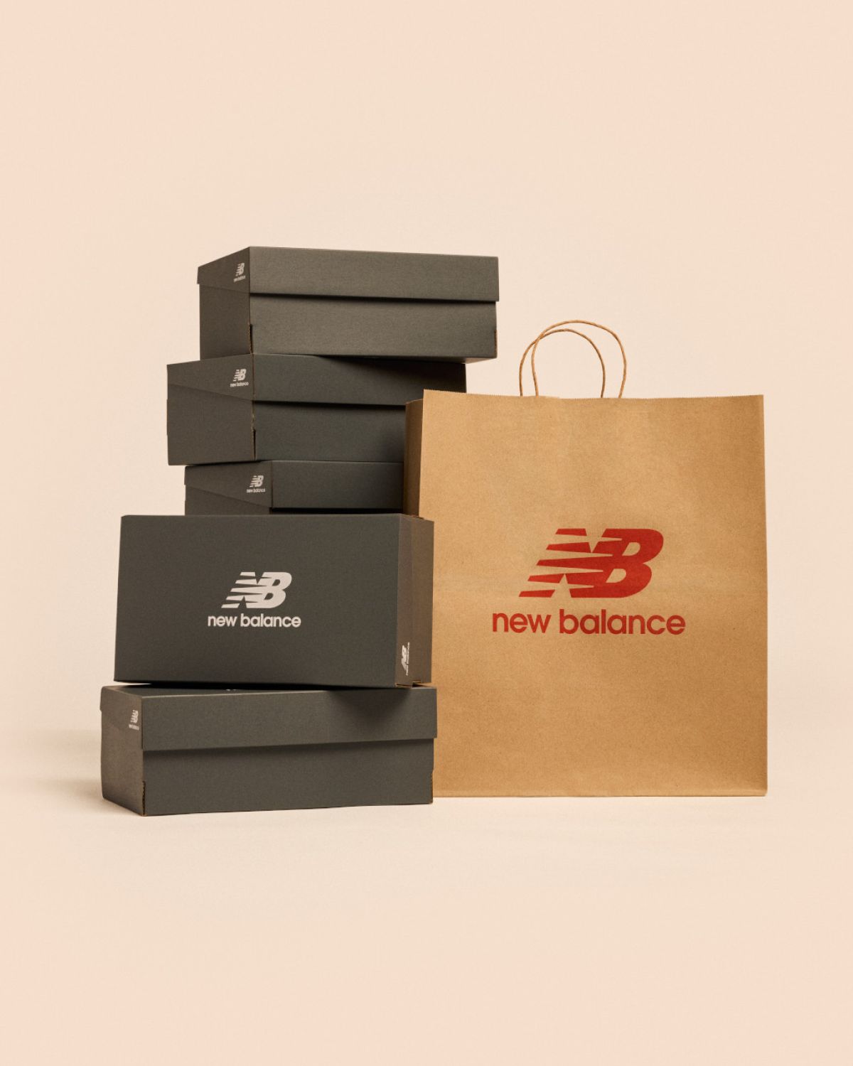Black shoeboxes are stacked in a small tower next to a brown paper shopping bag with the red NB New Balance icon and logo