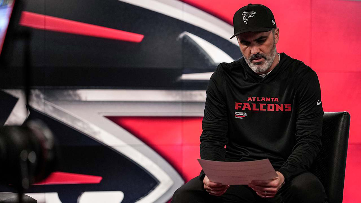 Kevin Stefanski seated holding papers in front of a large Falcons logo backdrop