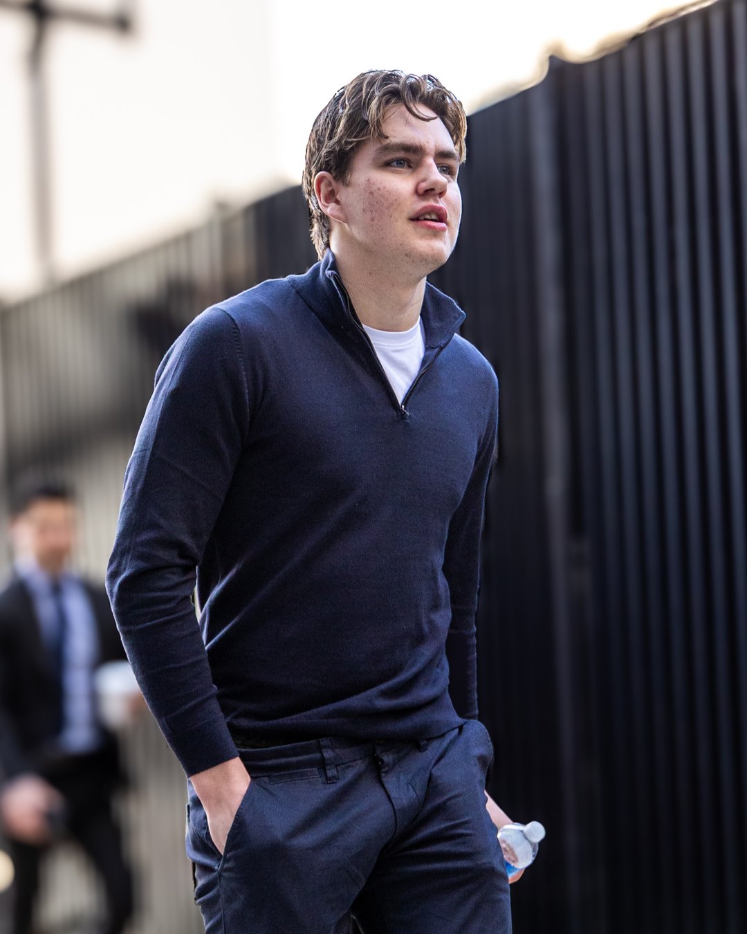 Young man walking outdoors, wearing dark clothes, holding a water bottle.