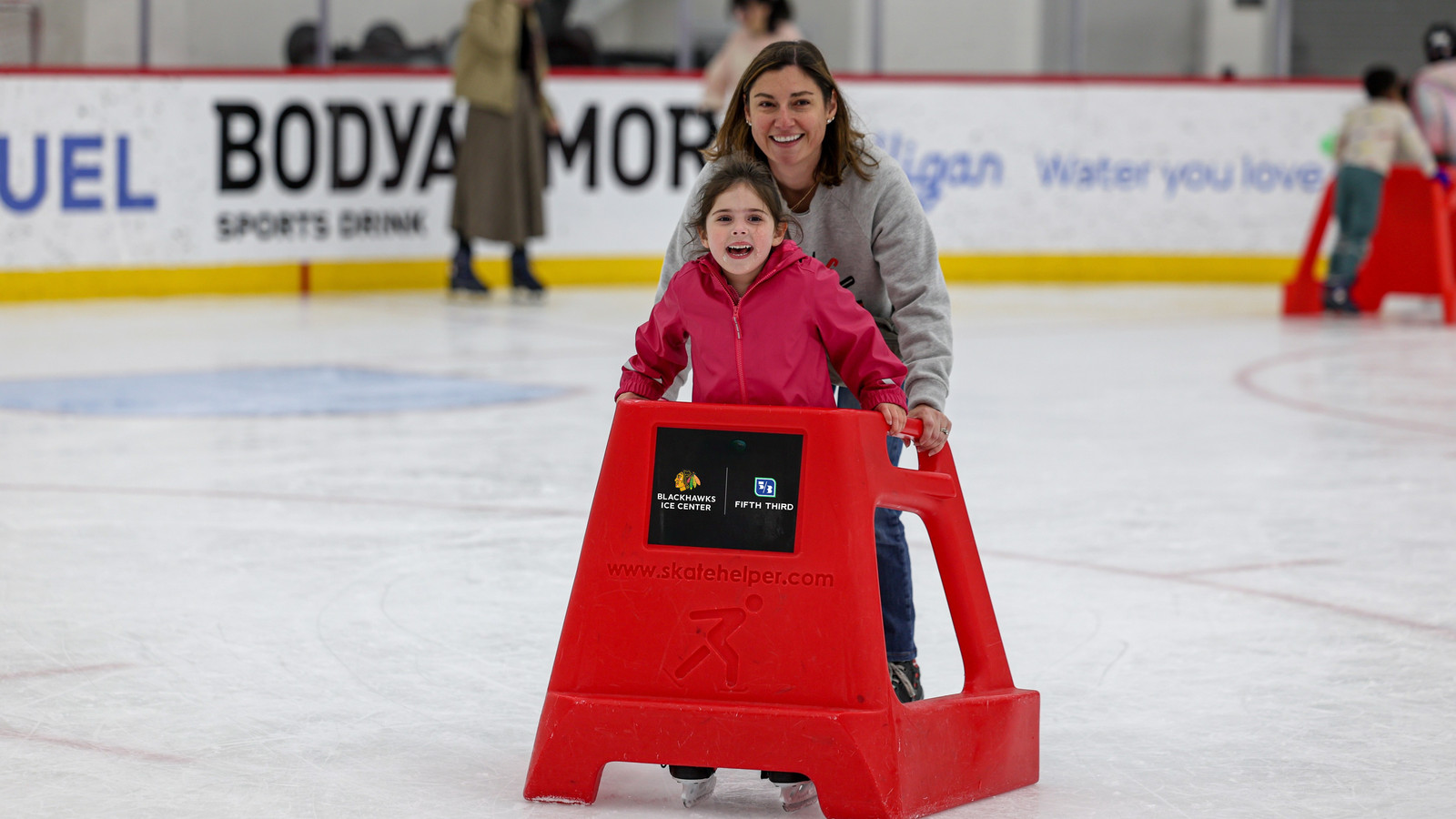 Woman and child ice skating with a red skating aid, both smiling.