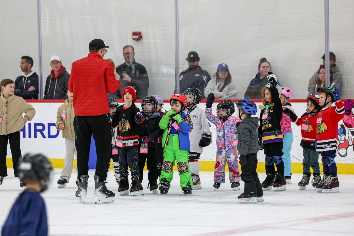Children on ice rink with adult instructor, some raising hands.
