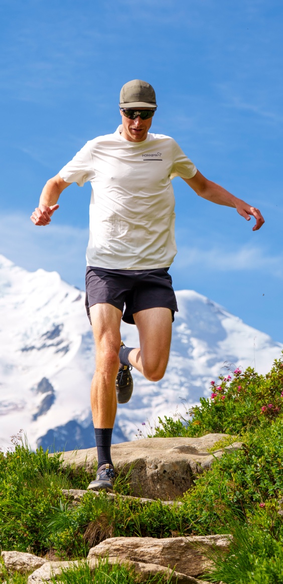 A man wearing a cap, sunglasses, a white t-shirt, and dark shorts trail runs down a steep, grassy path with snow-capped mountains in the background.